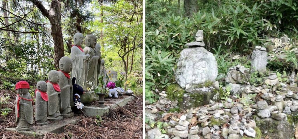 Jizo with bibs, toys, and flowers stand in a line in the forest. Stones are piled up around larger stones carved with Jizo.