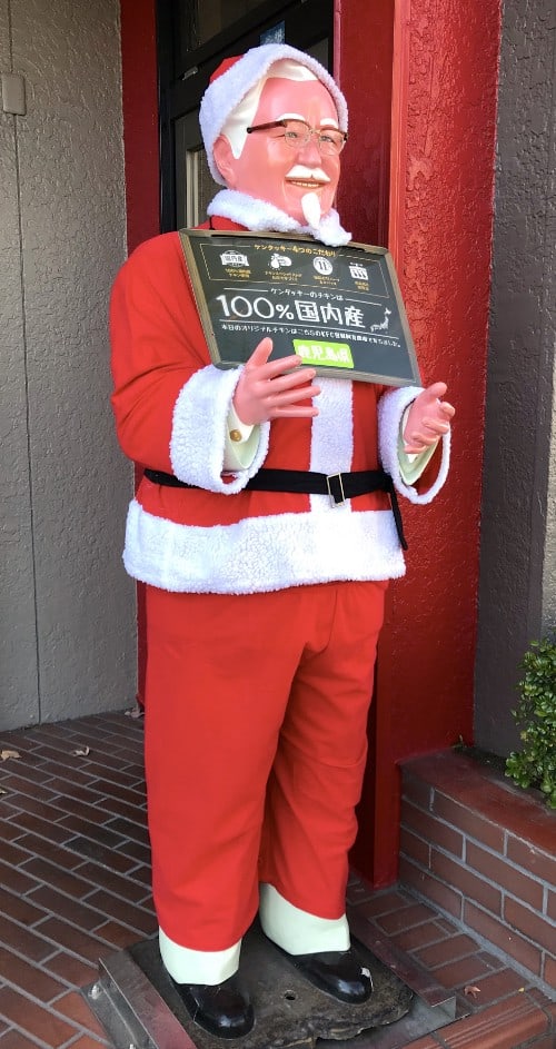 Kentucky Fried Chicken store with Colonel Sanders dressed as Santa. His sign reads, "Our chicken is locally sourced."