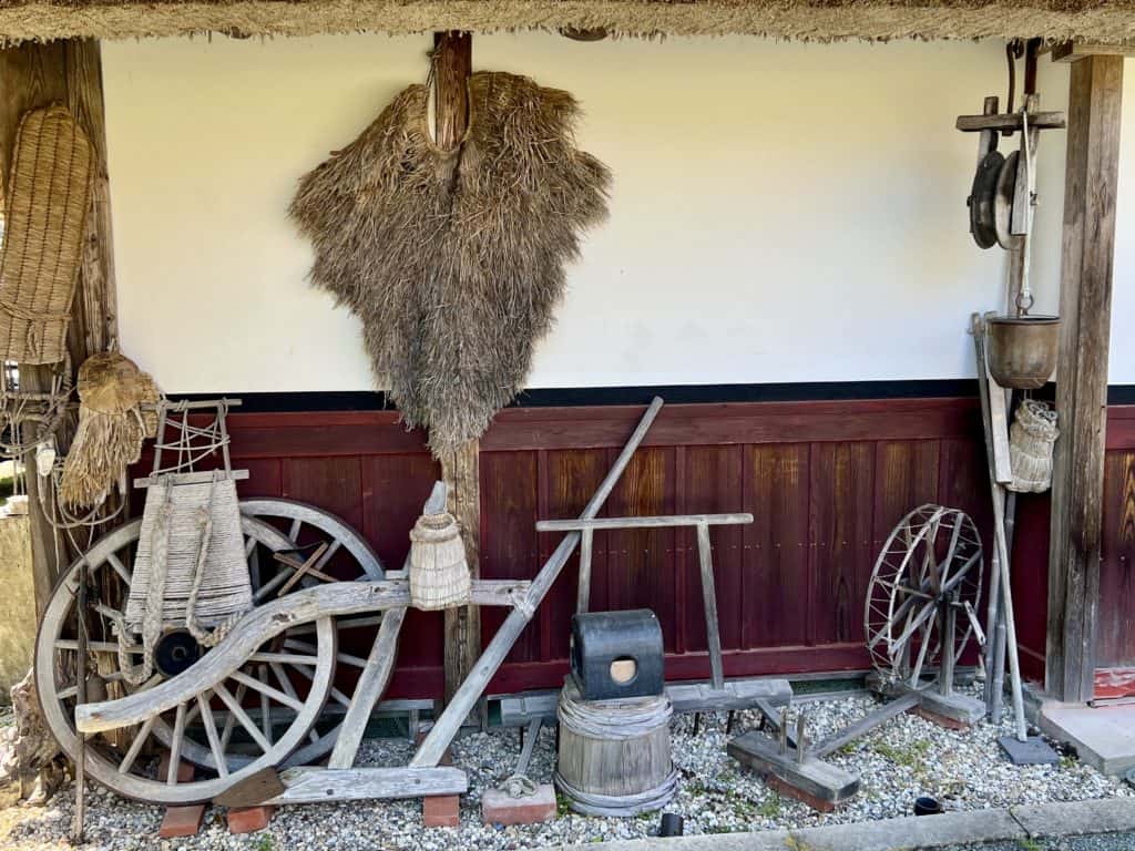 A thatched house with old farm equipment and a straw raincoat under the eaves.
