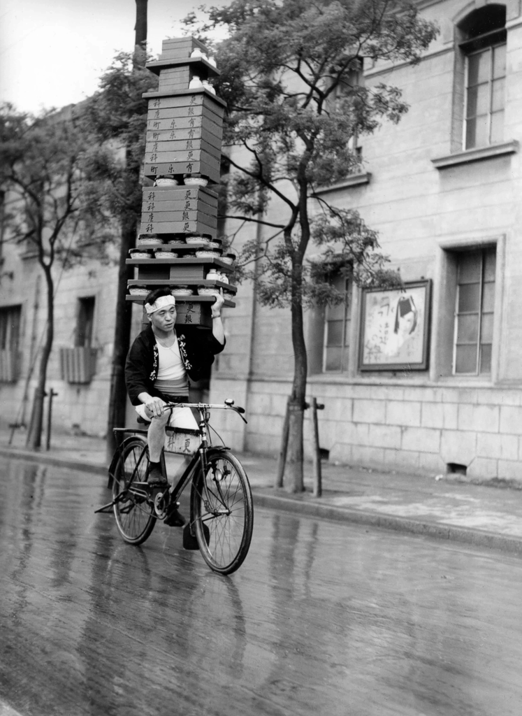 Man on bicycle with a tall stack of boxes and bowls on his shoulder.
